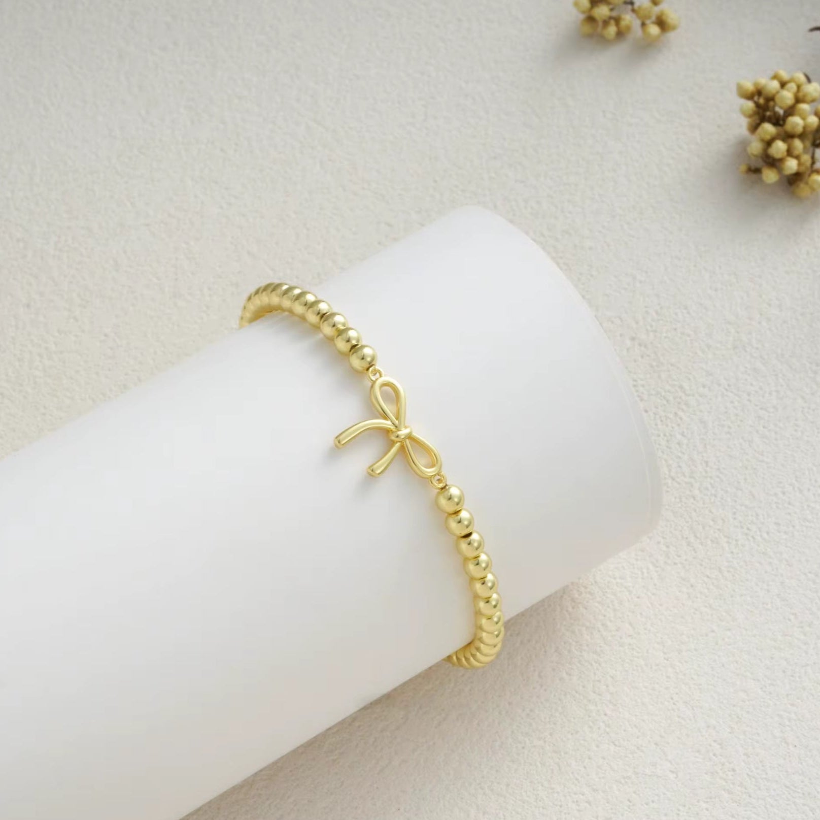 Gold Bow Bead Bracelet, on a white cylindrical object with a light beige background