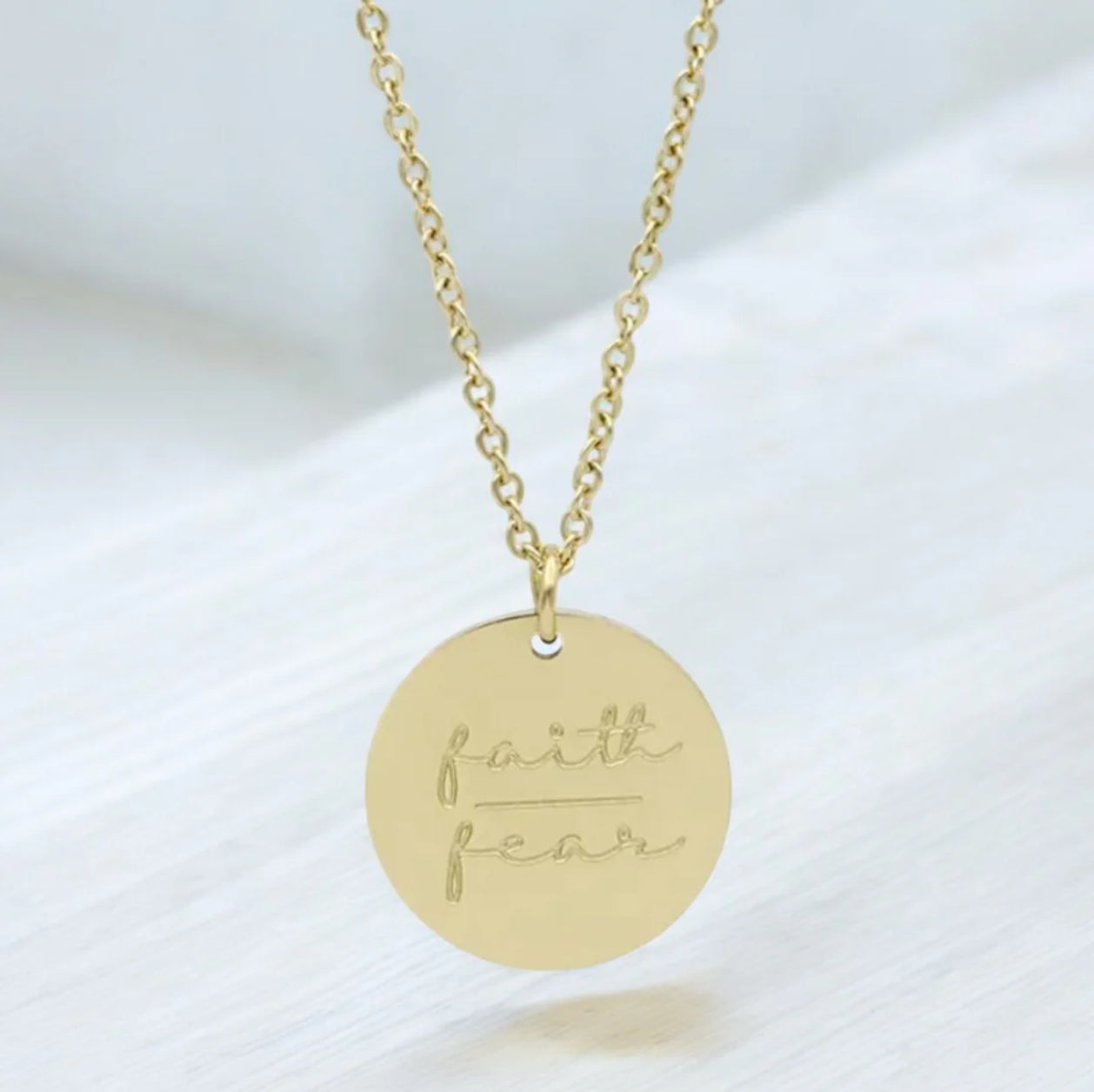 A gold necklace with a circular pendant engraved with the words 'faith over fear' on a chain link.