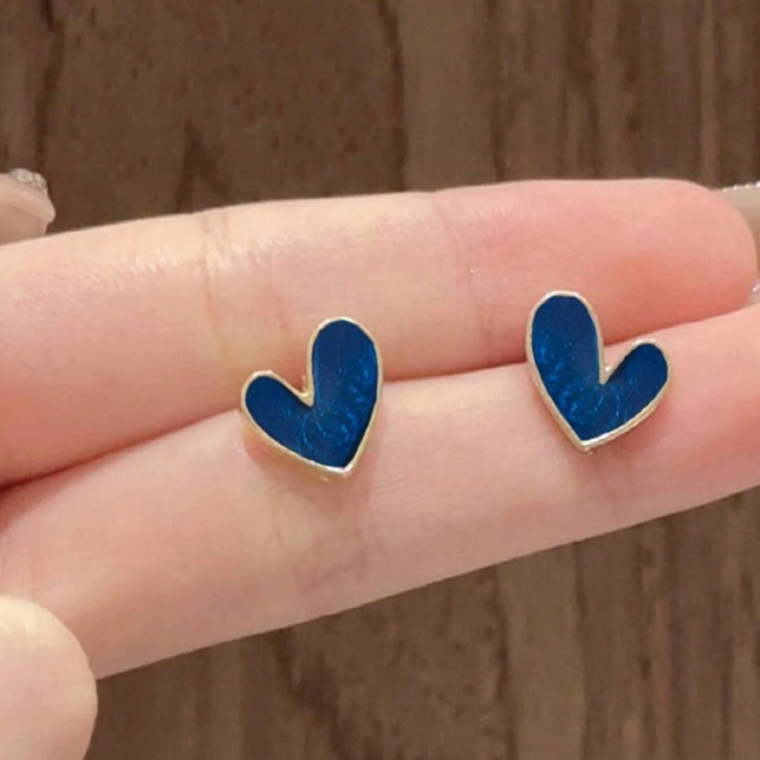 A pair of gold-tone stud earrings with a dark blue heart design, held between fingers against a wooden background.