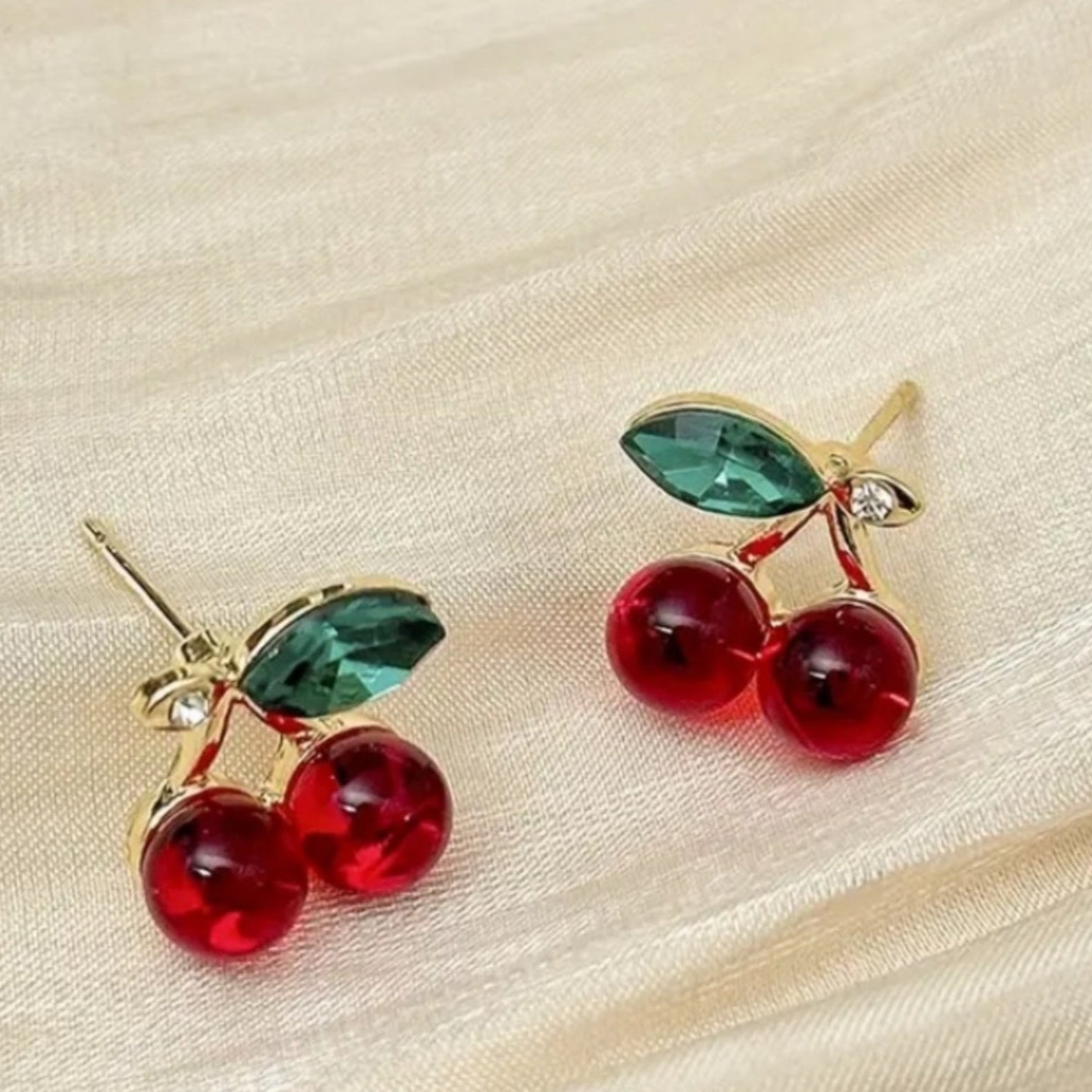 A pair of cherry-shaped earrings with glossy red color and green leaf accents, connected by gold-tone detailing with small zircon stones.