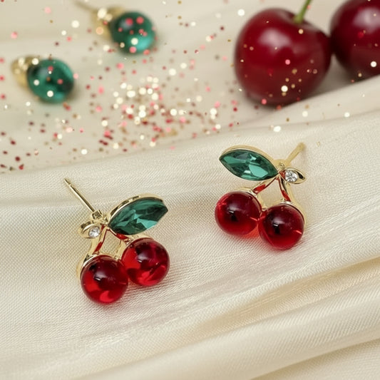 A pair of cherry-shaped earrings with glossy red color and green leaf accents, connected by gold-tone detailing with small zircon stones.