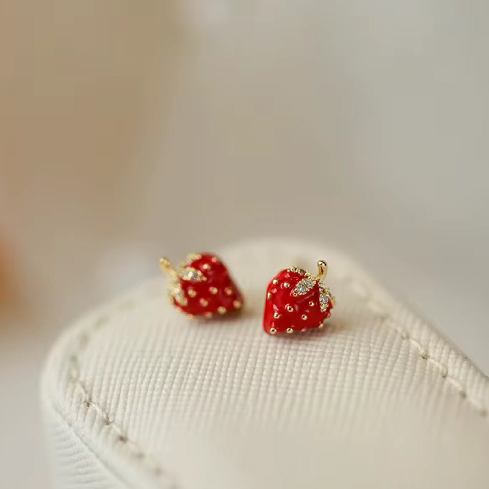 A pair of red strawberry-shaped earrings with gold-toned seeds and zircon-studded leaves displayed on a white stand.