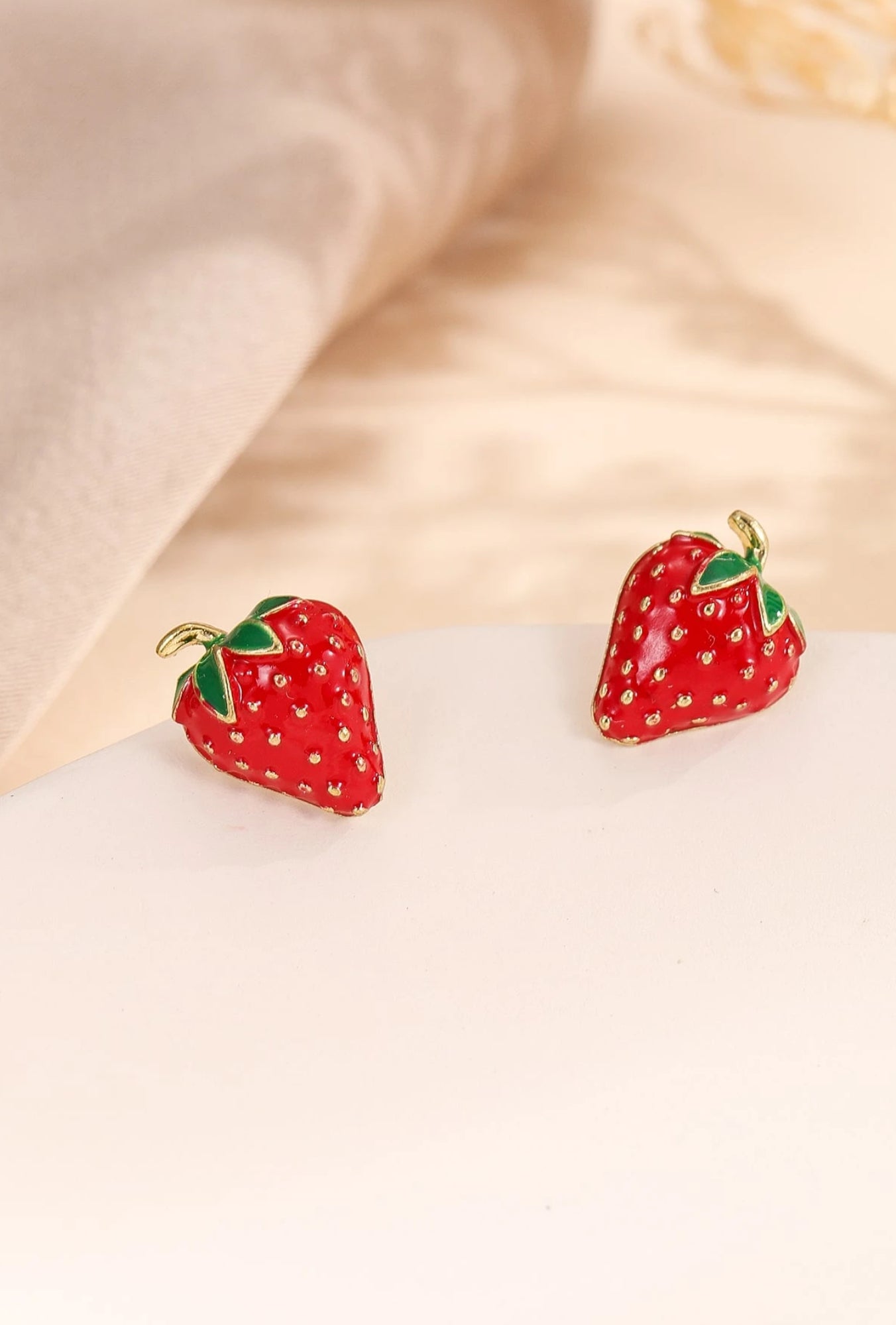 A pair of strawberry-shaped earrings with a glossy red finish, gold-toned seed details, and green leaf accents on a white surface.