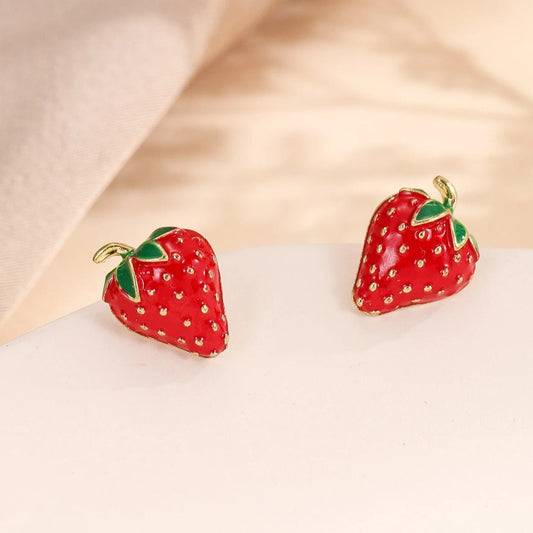 A pair of strawberry-shaped earrings with a glossy red finish, gold-toned seed details, and green leaf accents on a white surface.