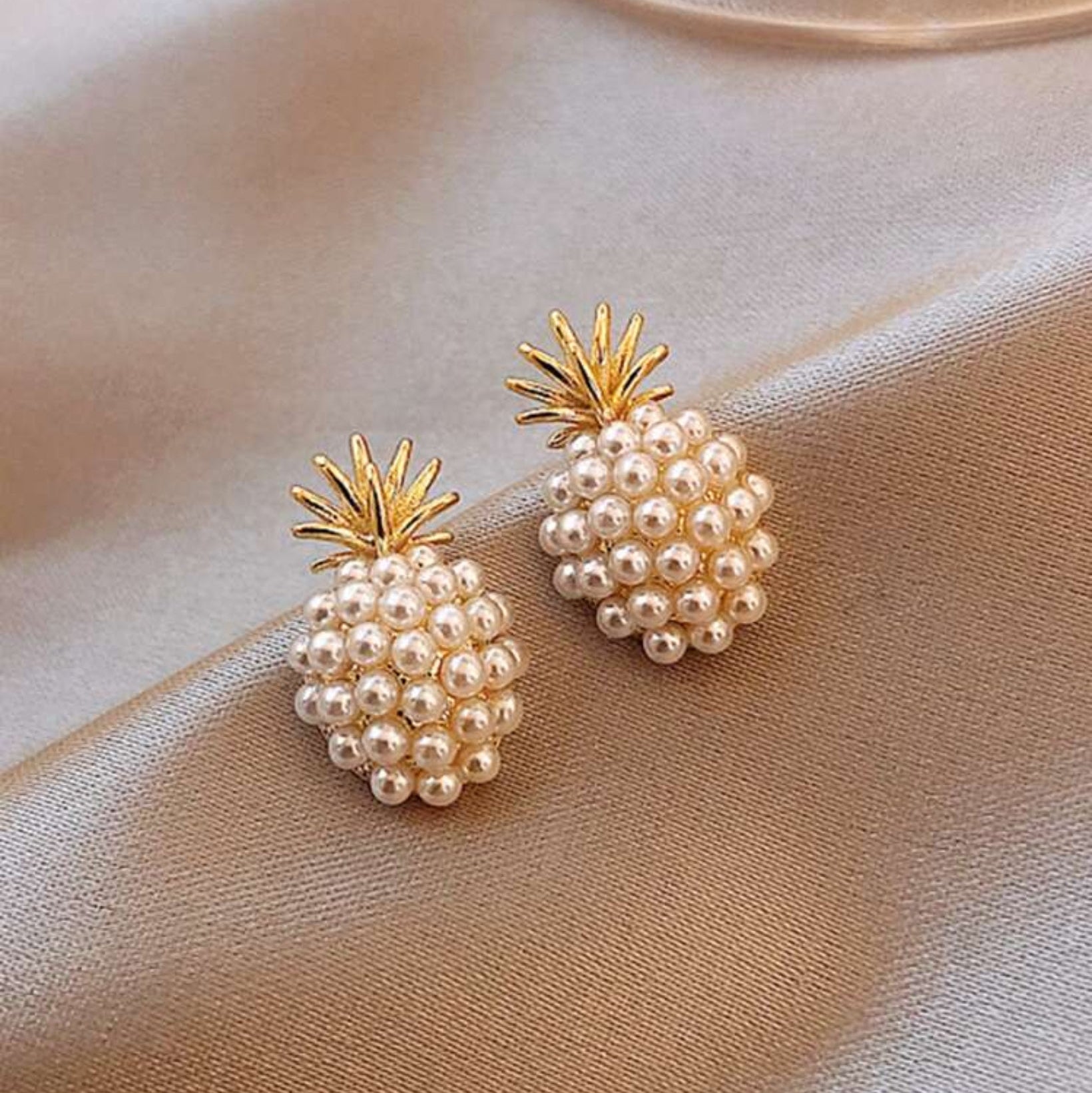 A pair of gold-tone pineapple-shaped earrings adorned with white imitation pearls, displayed on a beige fabric surface.