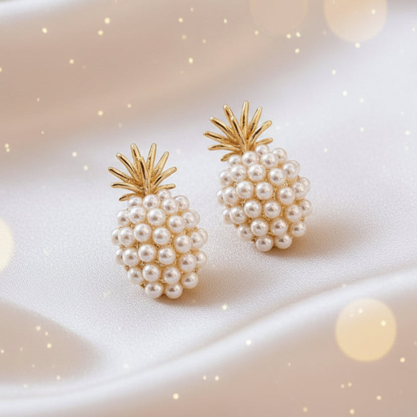 A pair of gold-tone pineapple-shaped earrings adorned with white imitation pearls, displayed on a beige fabric surface.
