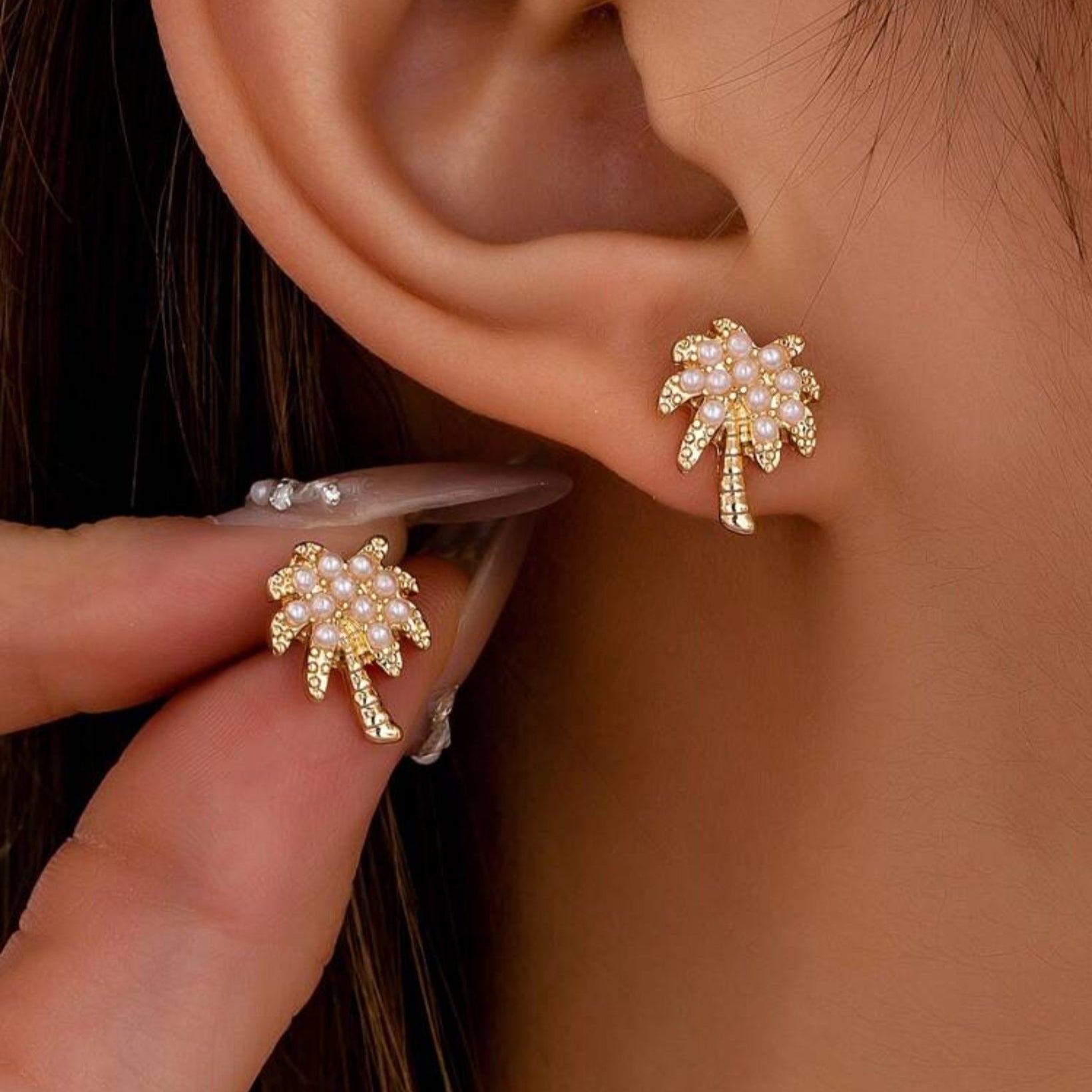 A pair of gold-tone palm tree earrings accented with white imitation pearls, worn on a person's ear.
