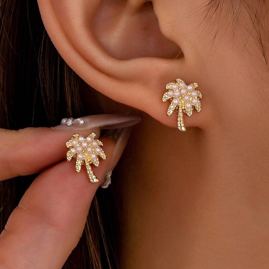 A pair of gold-tone palm tree earrings accented with white imitation pearls, worn on a person's ear.
