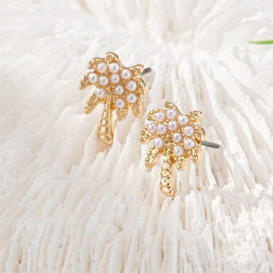 A pair of gold-tone palm tree earrings accented with white imitation pearls, displayed on a white textured surface.