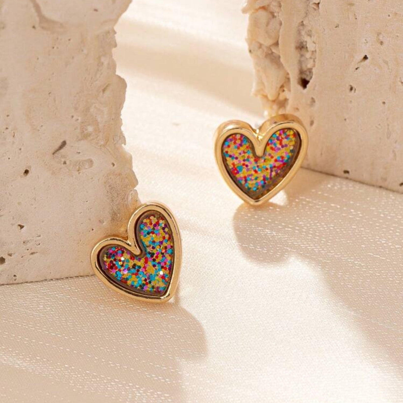 A pair of gold-tone heart-shaped stud earrings filled with multicolored confetti glitter, placed on a textured beige surface.