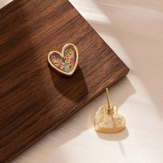 Gold-tone heart-shaped stud earrings filled with confetti glitter, displayed on a wooden surface.