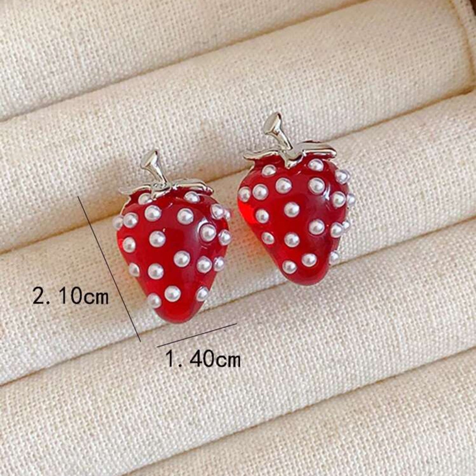 Red strawberry-shaped earrings with pearl-like embellishments on a beige fabric background.