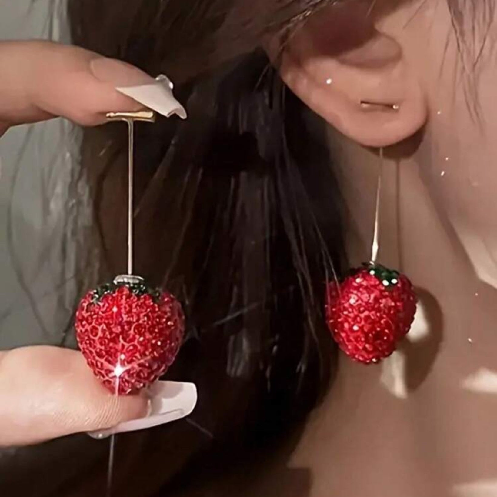 Red strawberry-shaped earrings being held up to a person's ear.