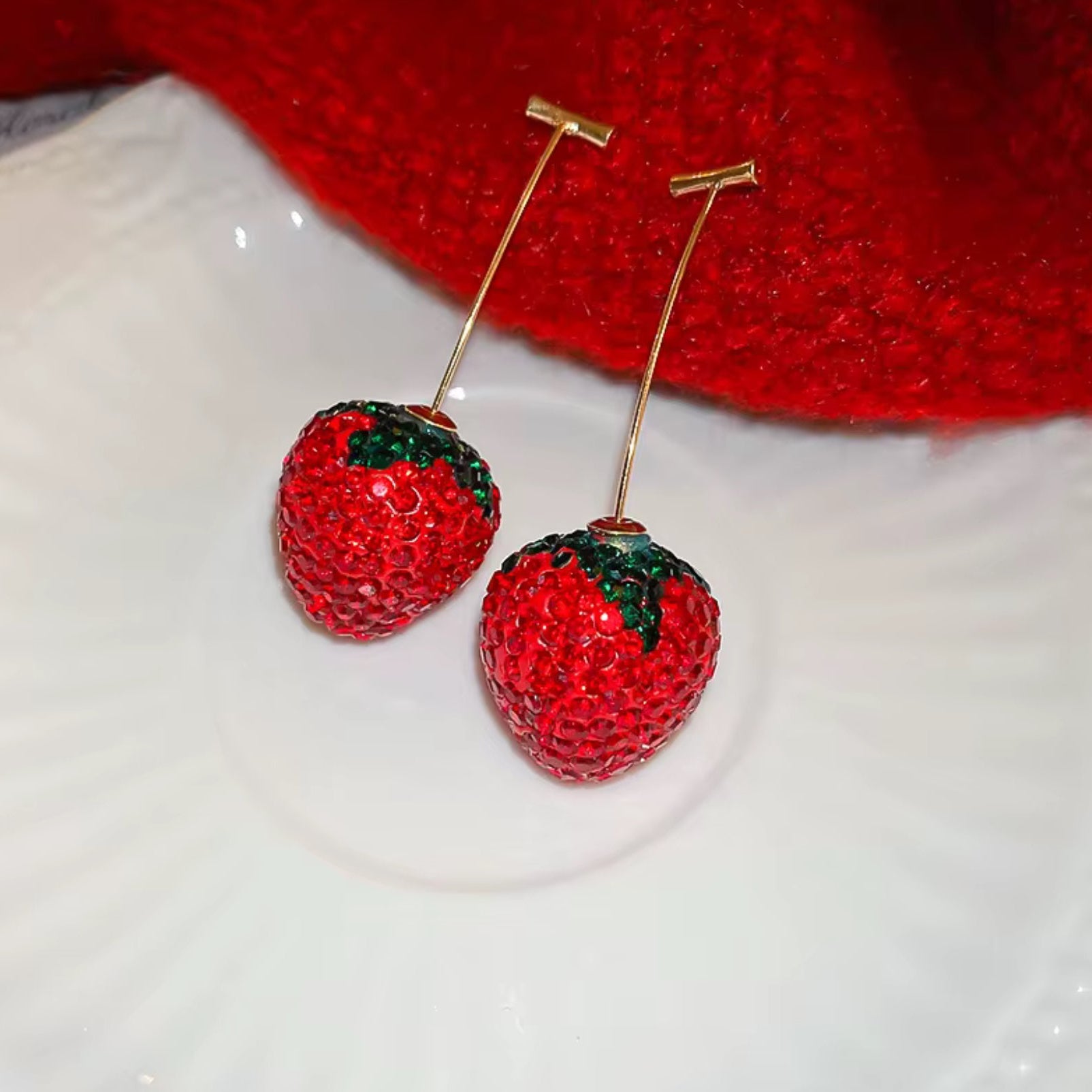 Red strawberry-shaped earrings on a white surface with a red fabric background