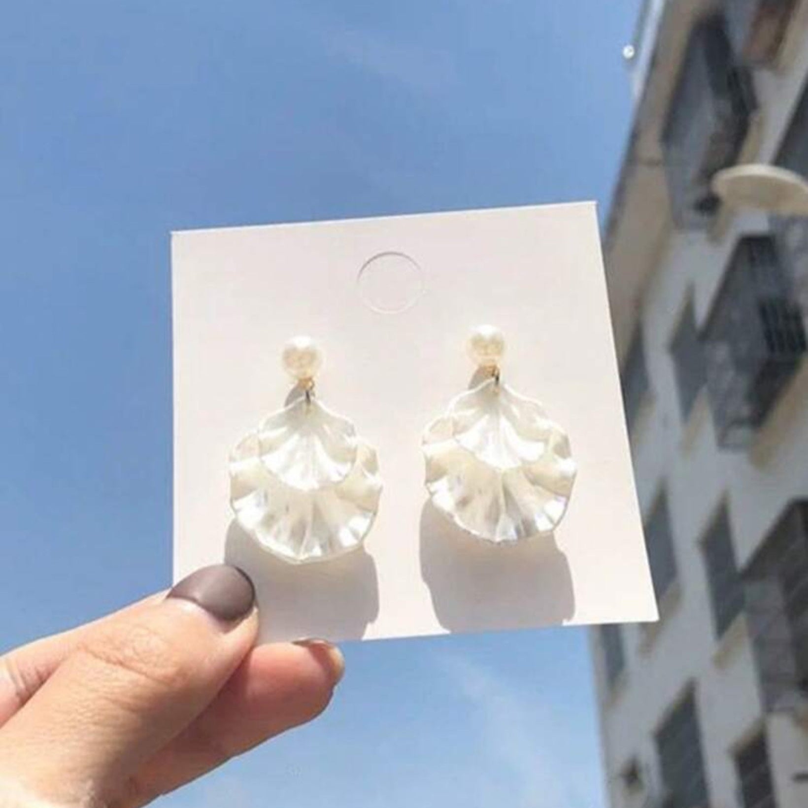 Pearl-like earrings on a card held against a blue sky with buildings in the background