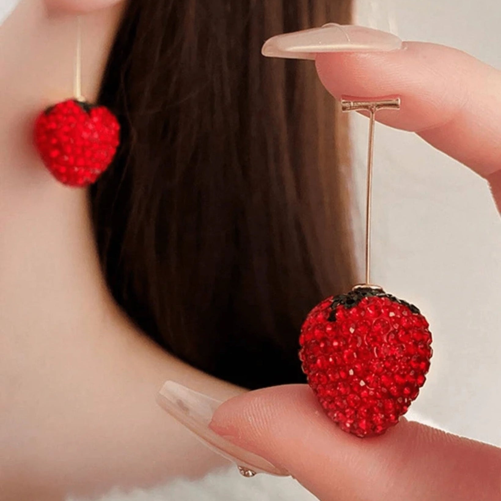Red strawberry-shaped earrings held by a hand against a blurred background