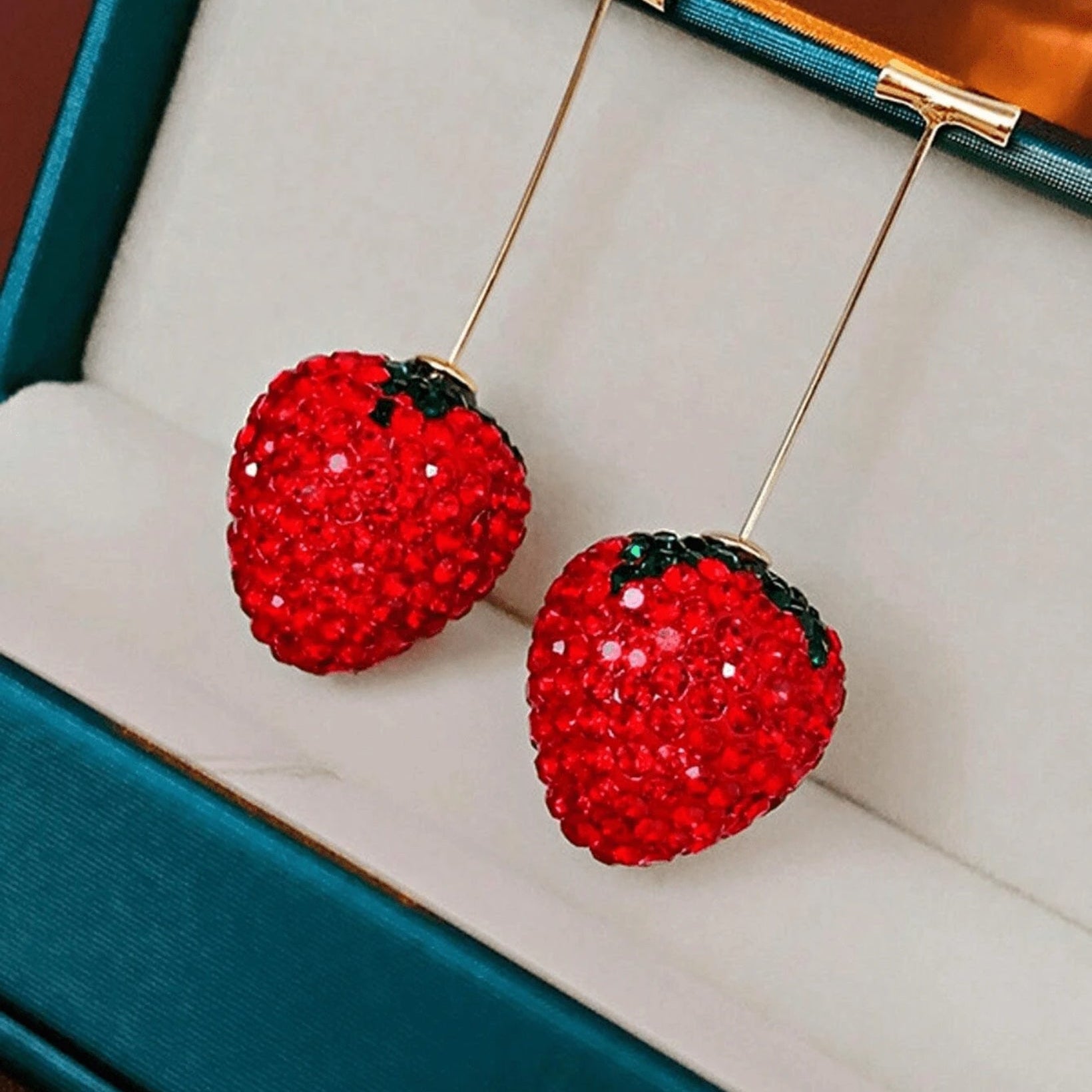 Red strawberry-shaped earrings with gold hooks on a white cushioned jewelry box.