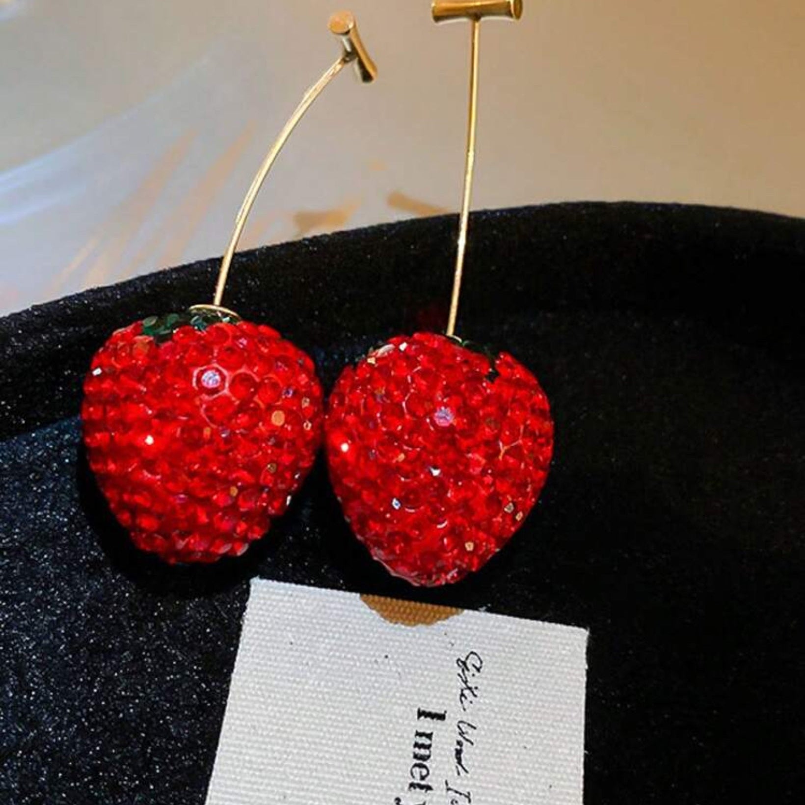 Red cherry-shaped earrings with gold hooks on a black stand with a white card.