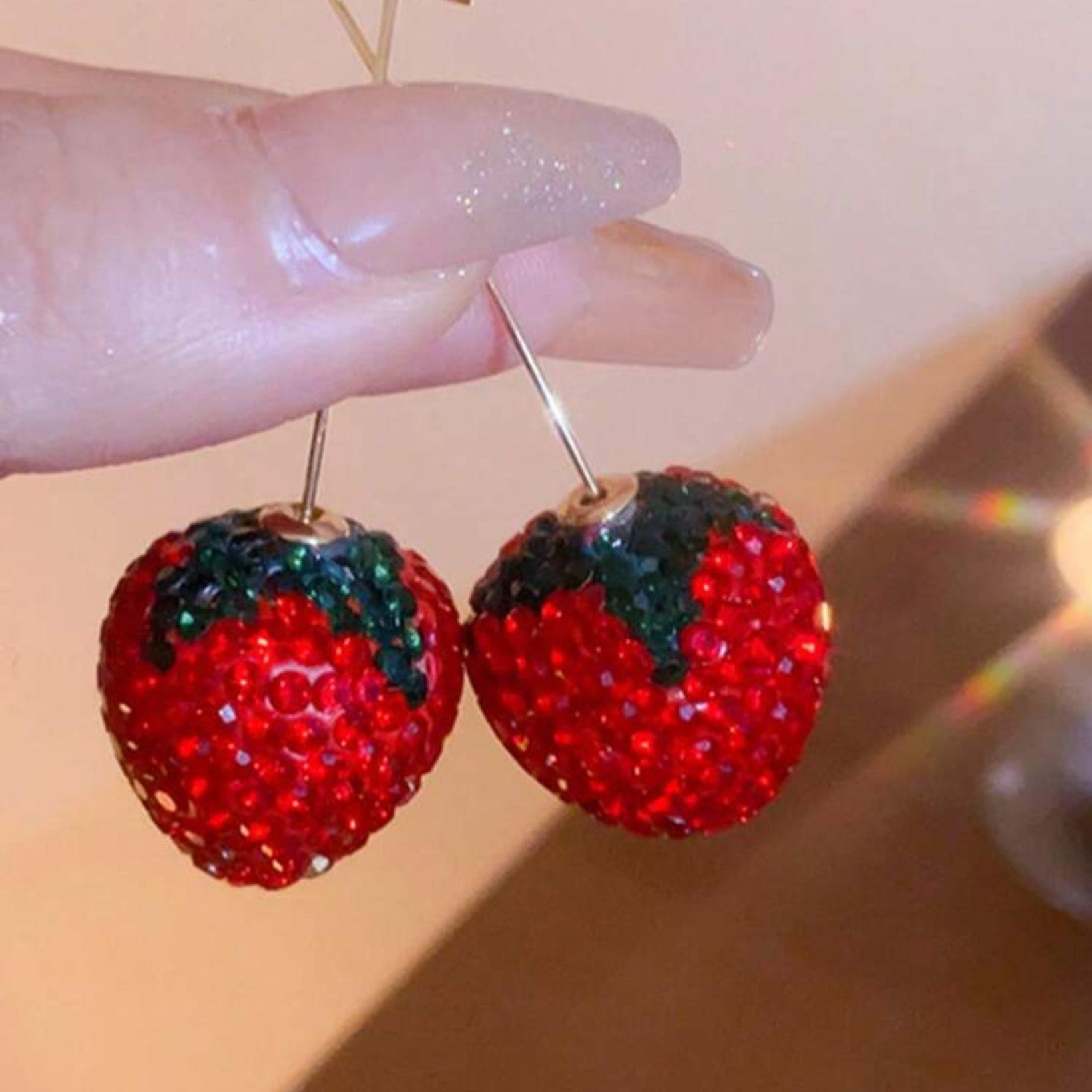 Red strawberry-shaped earrings with green leaves held by a hand against a neutral background