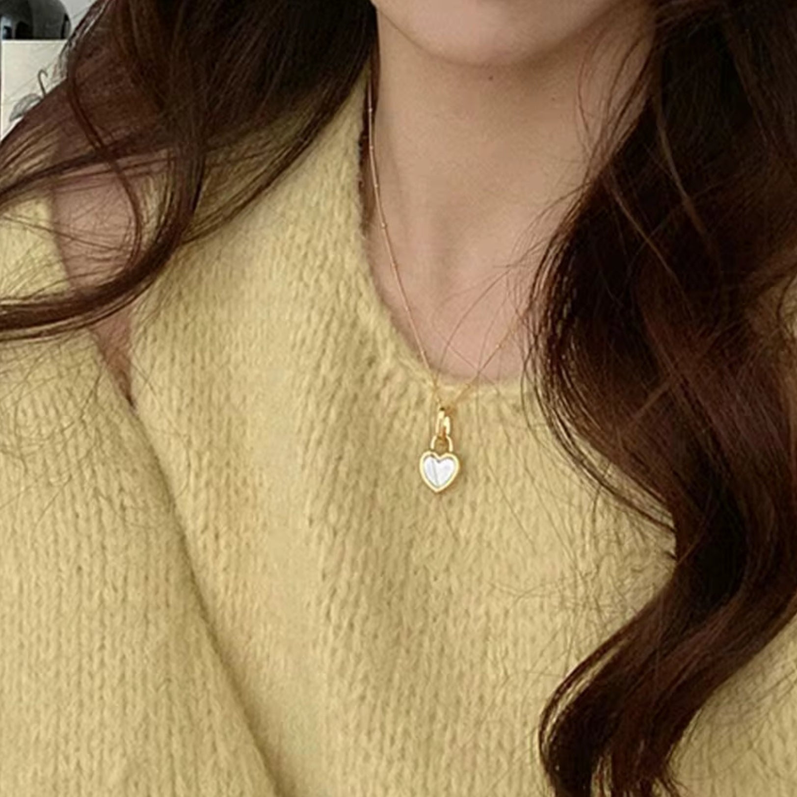 Close-up of a person wearing a gold necklace with a heart-shaped pendant, against a neutral background.