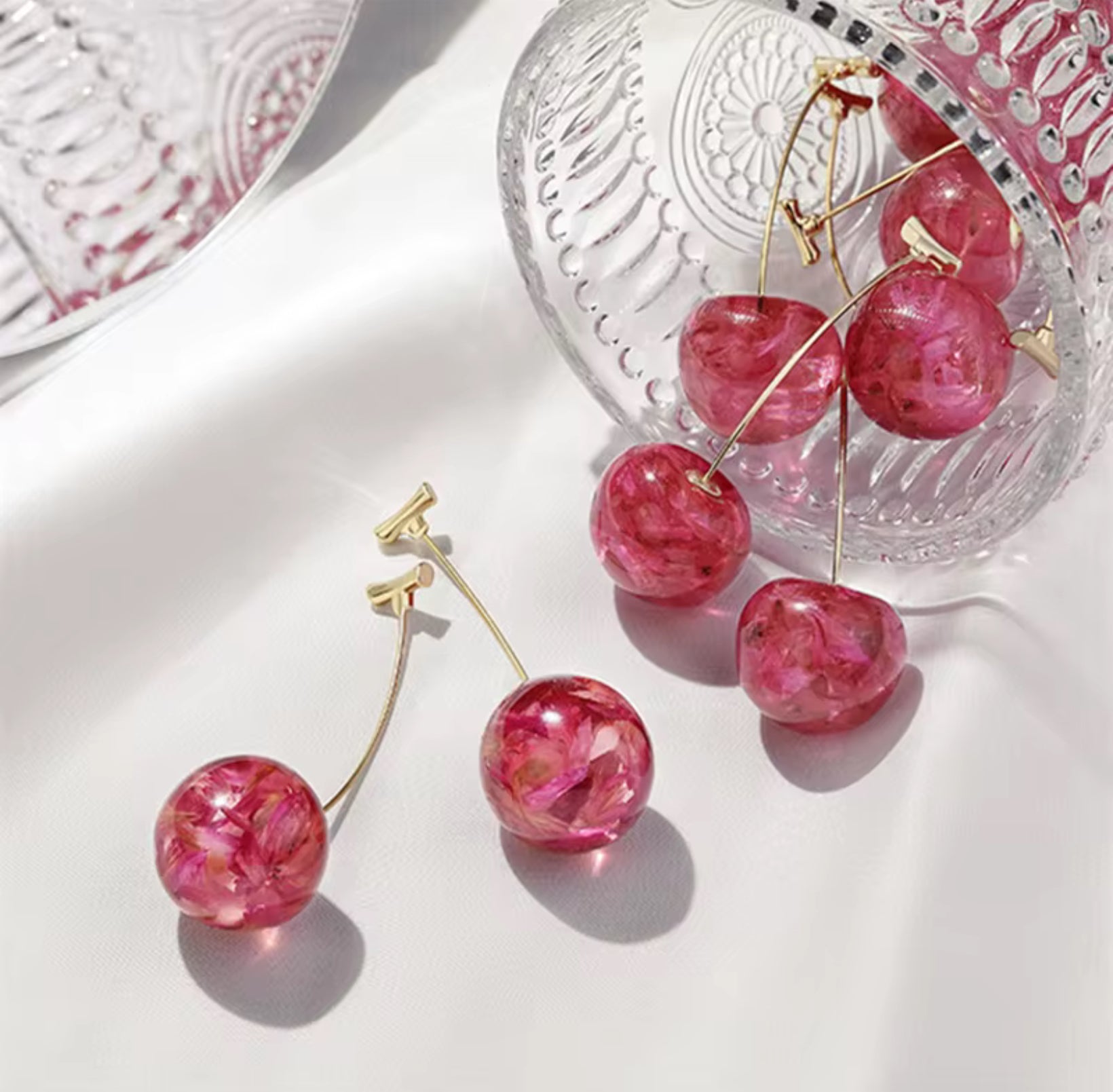 Pink marbled cherry-shaped earrings on a white surface with a glass dish in the background.