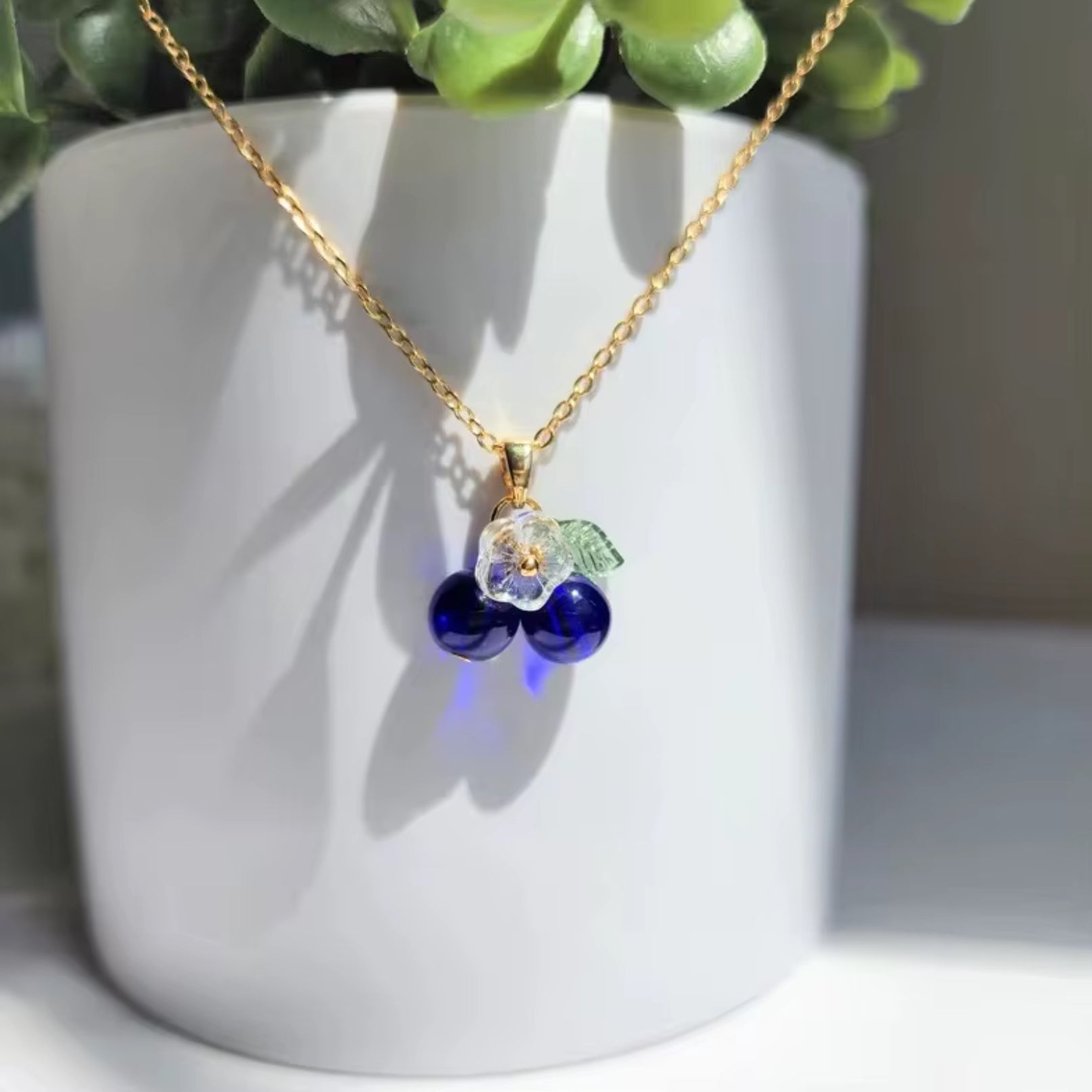 Necklace with blue beads and a leaf charm on a white surface with a plant in the background