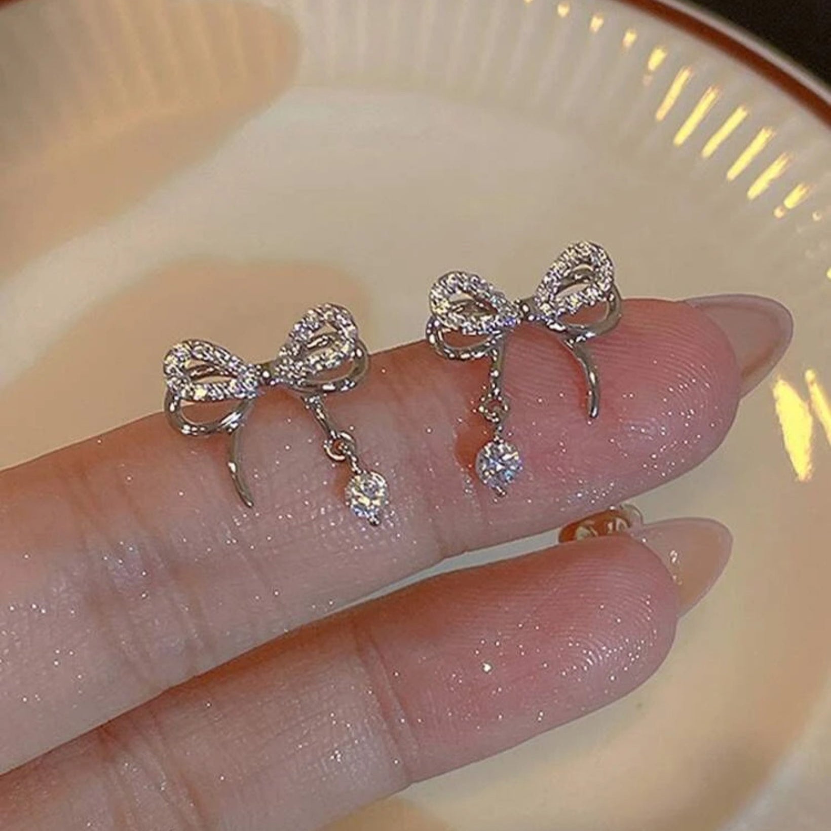 Silver bow-shaped earrings with dangling crystals on a hand against a neutral background