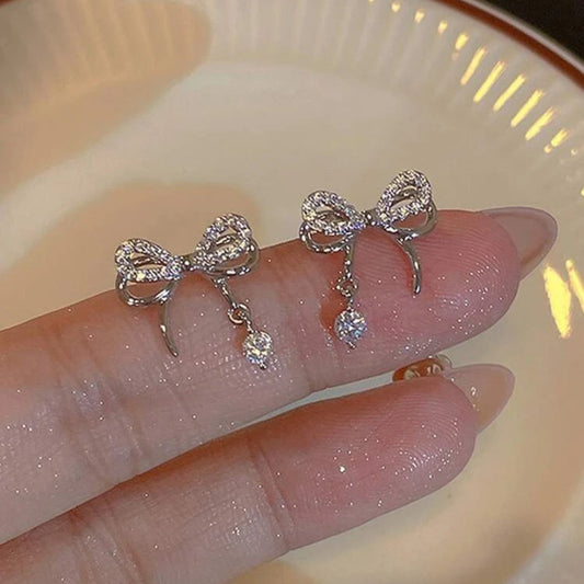 Silver bow-shaped earrings with dangling crystals on a hand against a neutral background