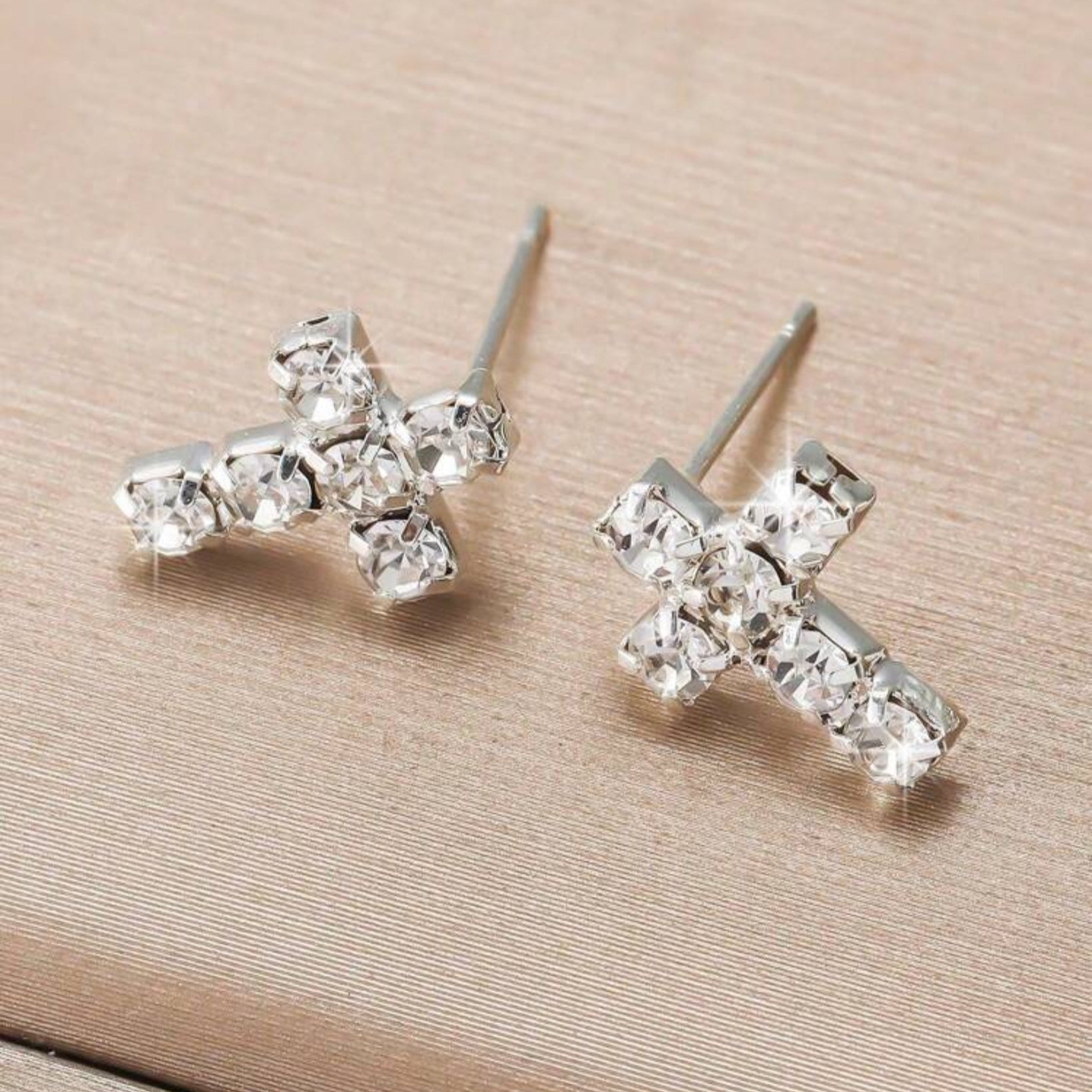 Silver cross-shaped earrings with clear gemstones on a beige surface