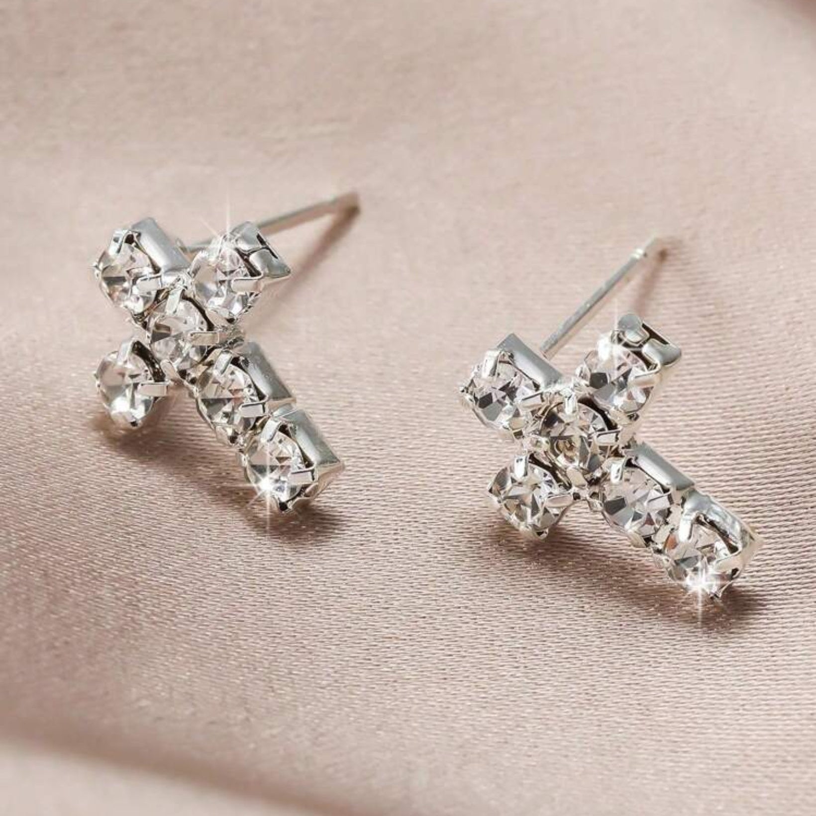 Silver cross-shaped earrings with clear gemstones on a beige fabric background