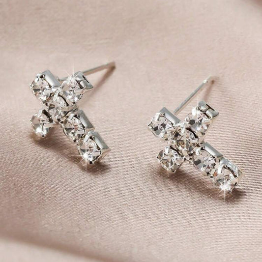 Silver cross-shaped earrings with clear gemstones on a beige fabric background