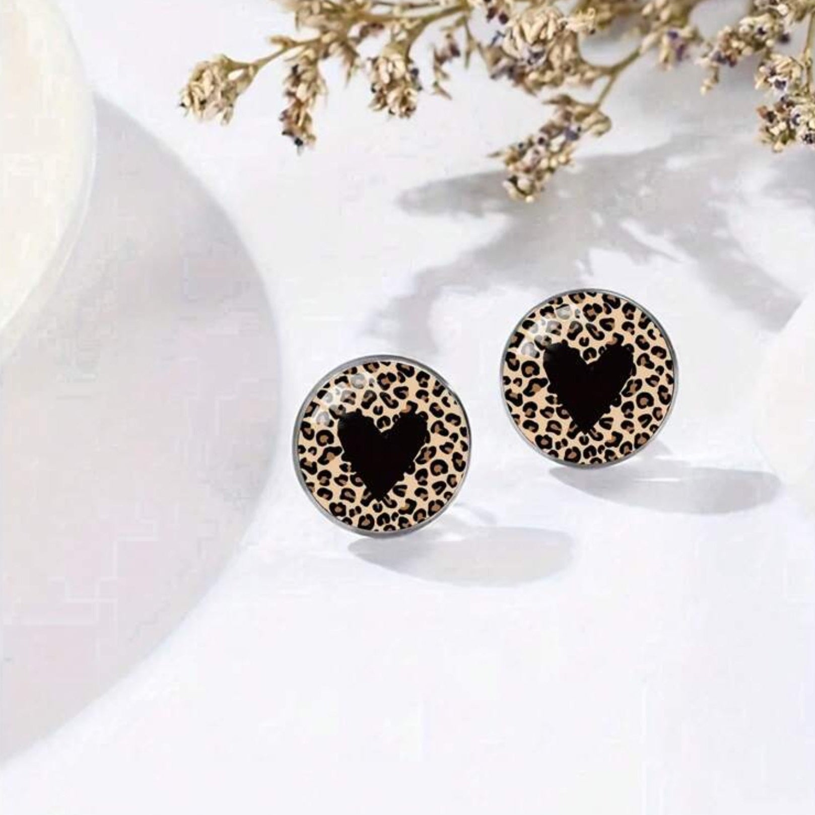 Leopard print earrings with black heart design on a white surface with dried flowers.