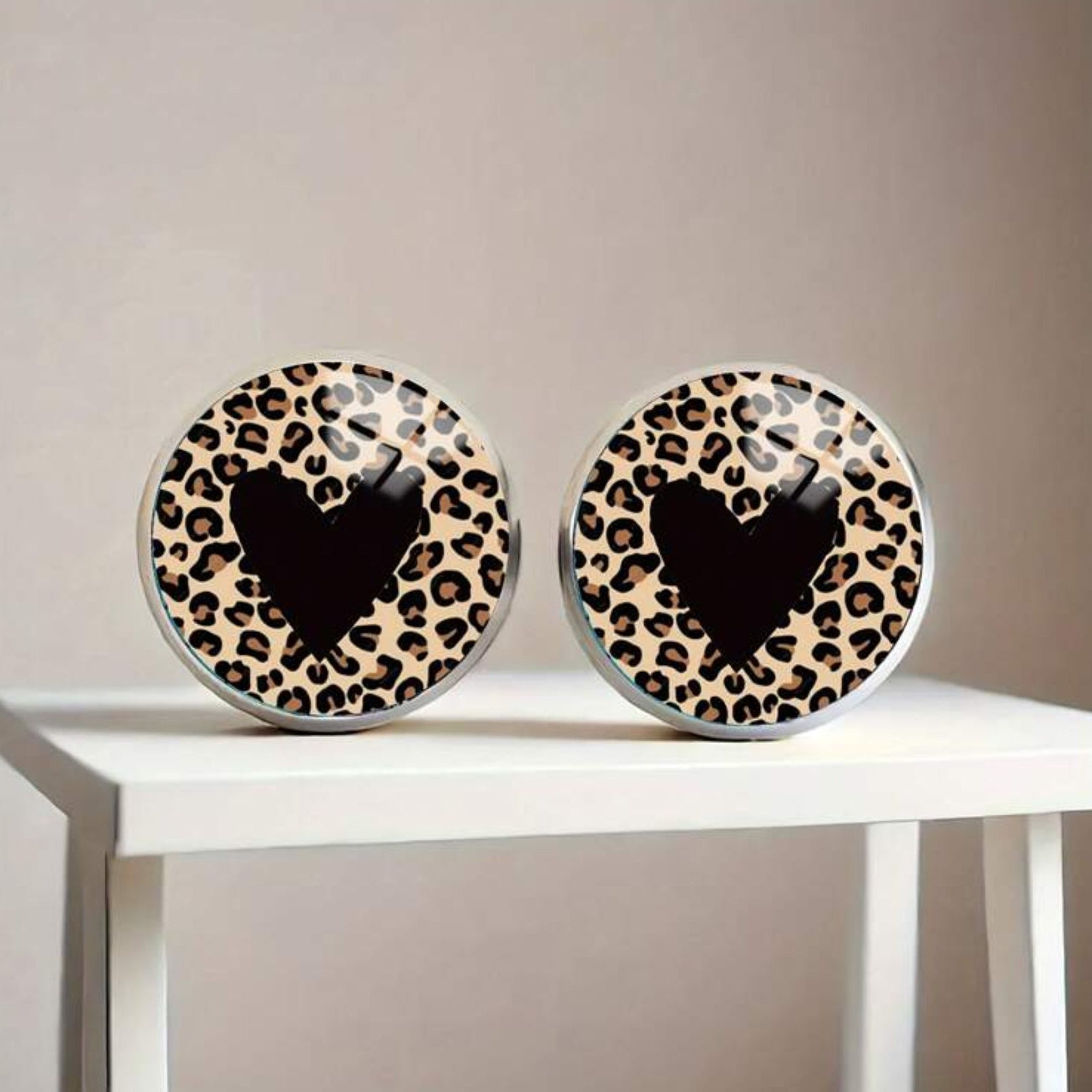 Two round earrings with leopard print pattern and black heart in the center on a white surface.