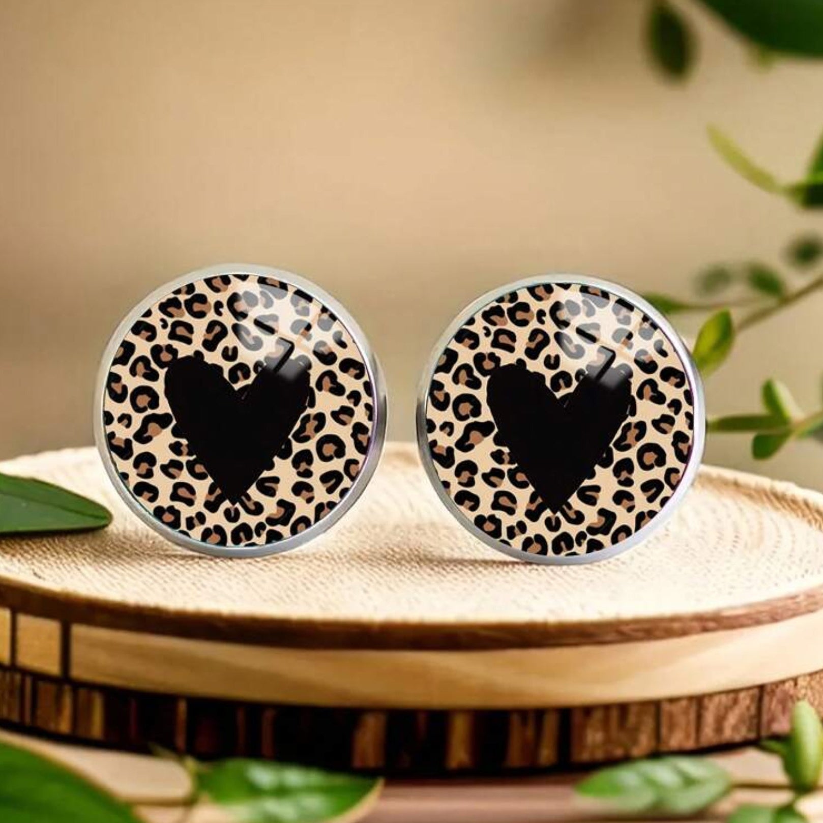 Pair of round earrings with leopard print and black heart design on a wooden surface.