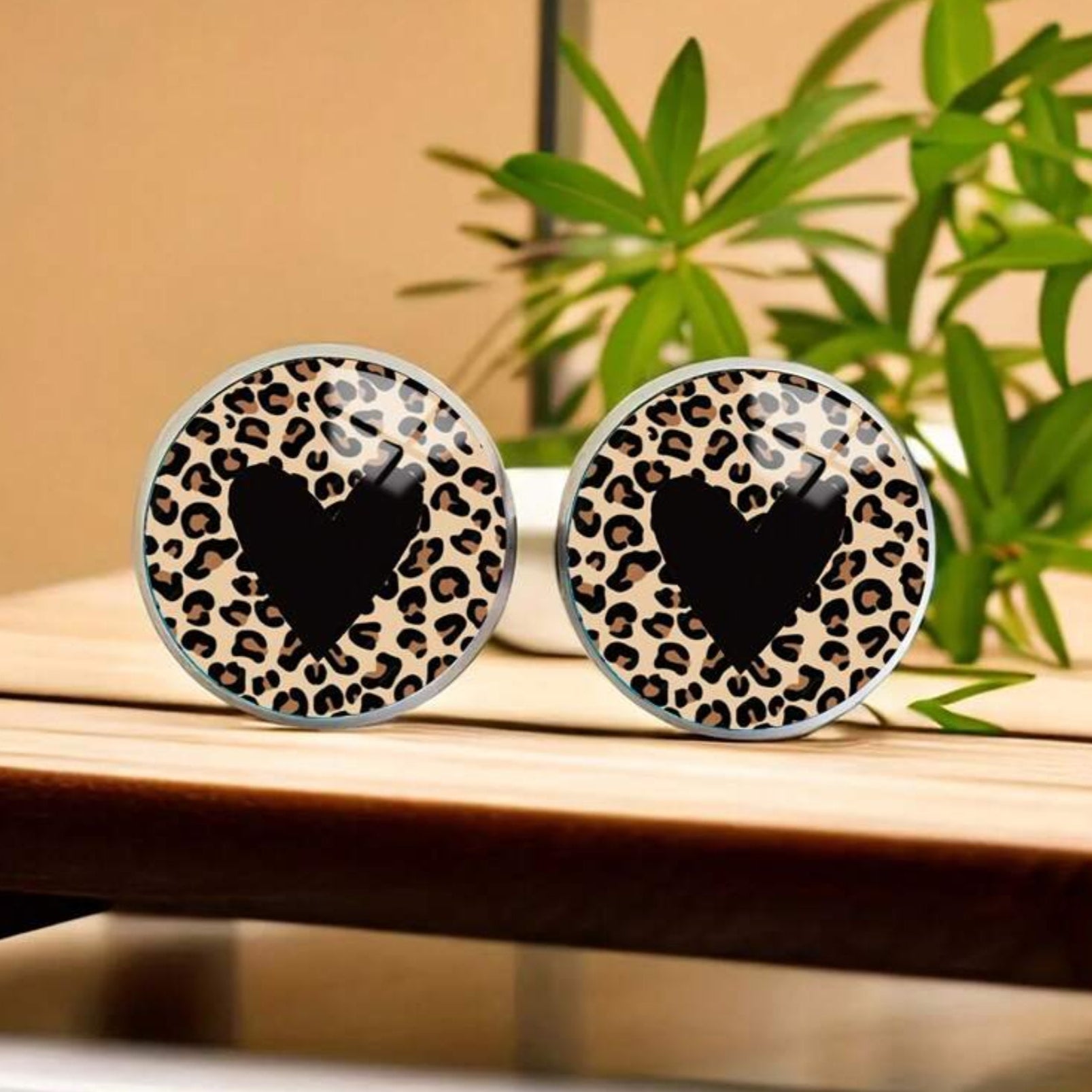 Leopard print earrings with a black heart design on a wooden surface with a plant in the background.