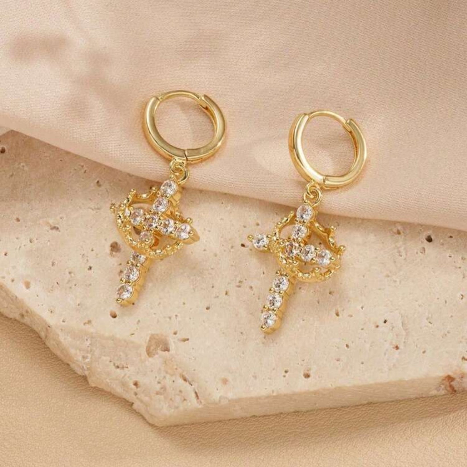 Gold Cross Crown earrings with cubic zirconia stones with intricate designs on a beige stone surface