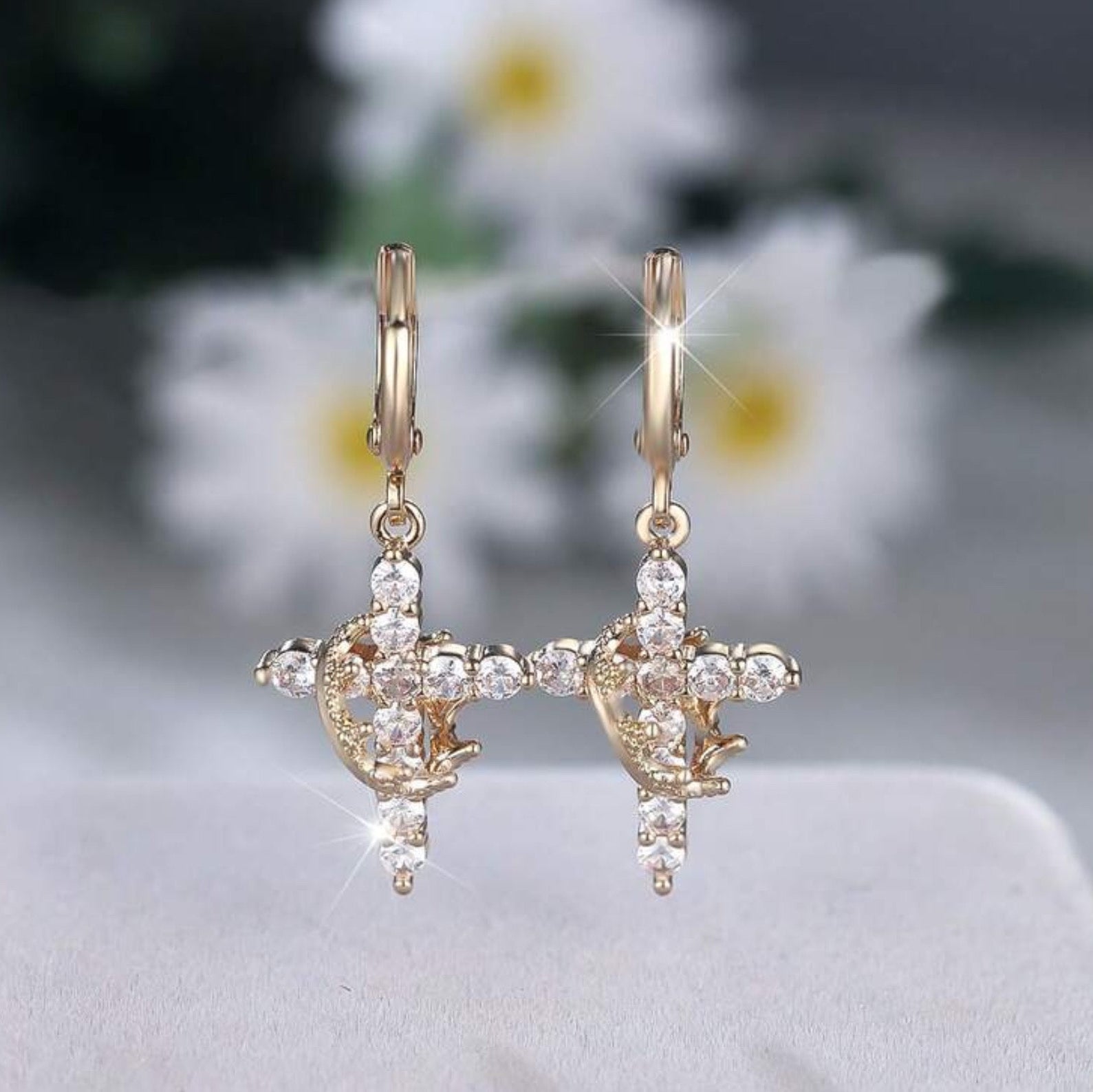 Gold Cross Crown earrings with cubic zirconia stones on a light gray background with blurred flowers in the background