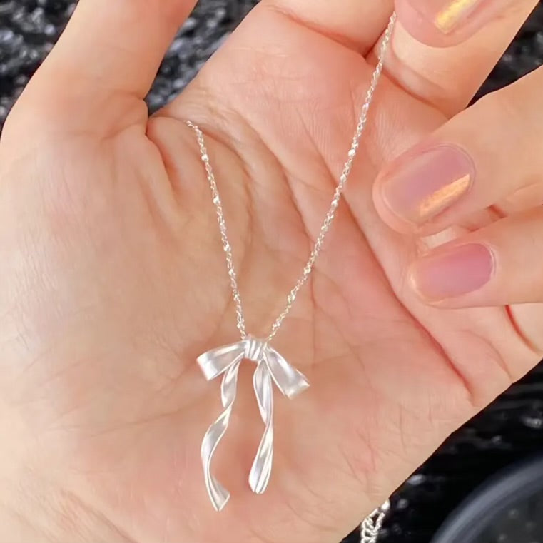 Silver Ribbon Bow Necklace with a bow pendant held in a hand against a black background