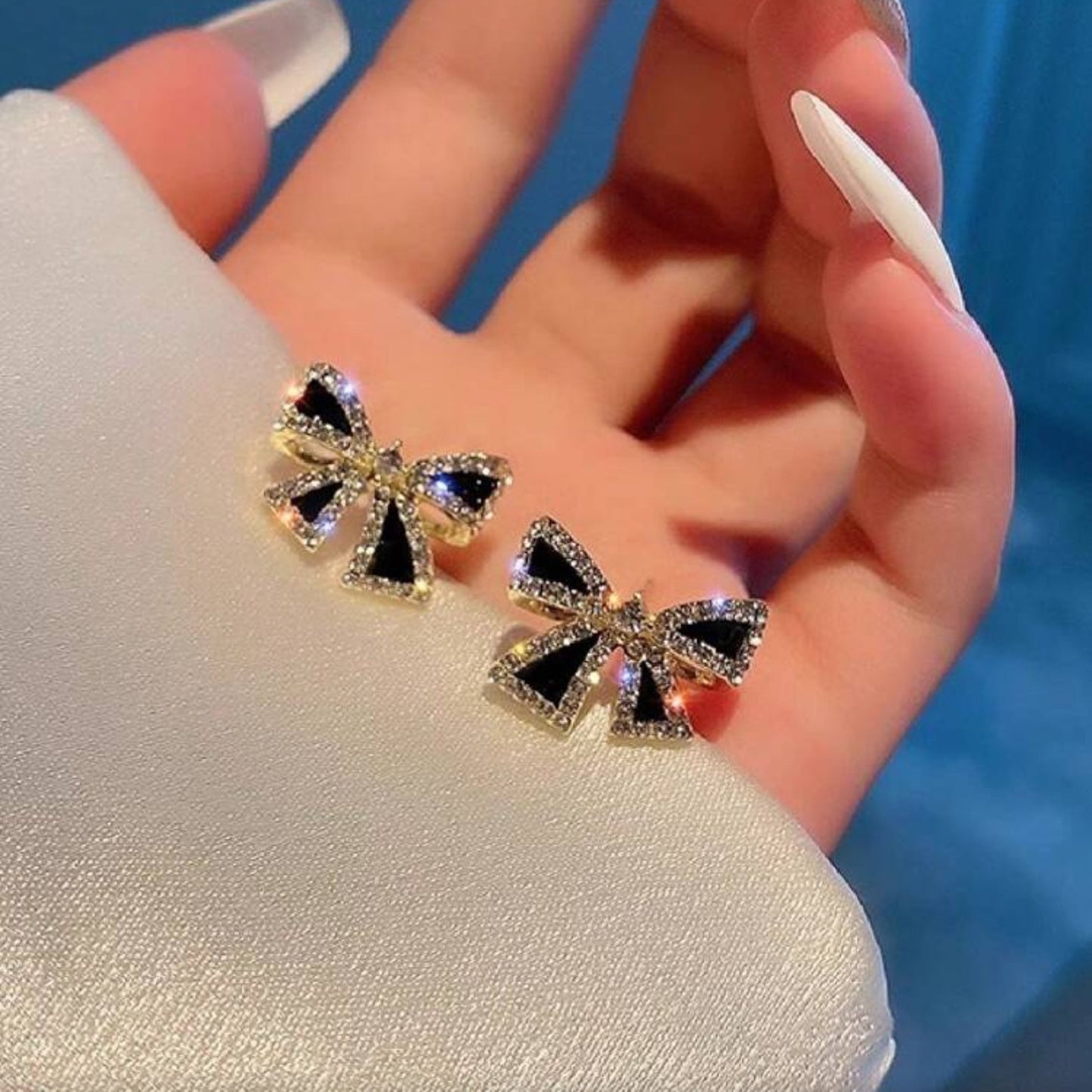 Black Bow Rhinestone Earrings held between fingers against a blue background