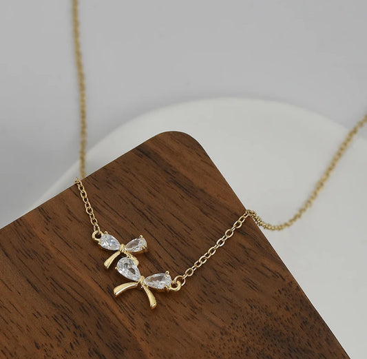 Zircon Bow Necklace featuring a sparkling bow pendant on an adjustable 17-19" chain, wood background.
