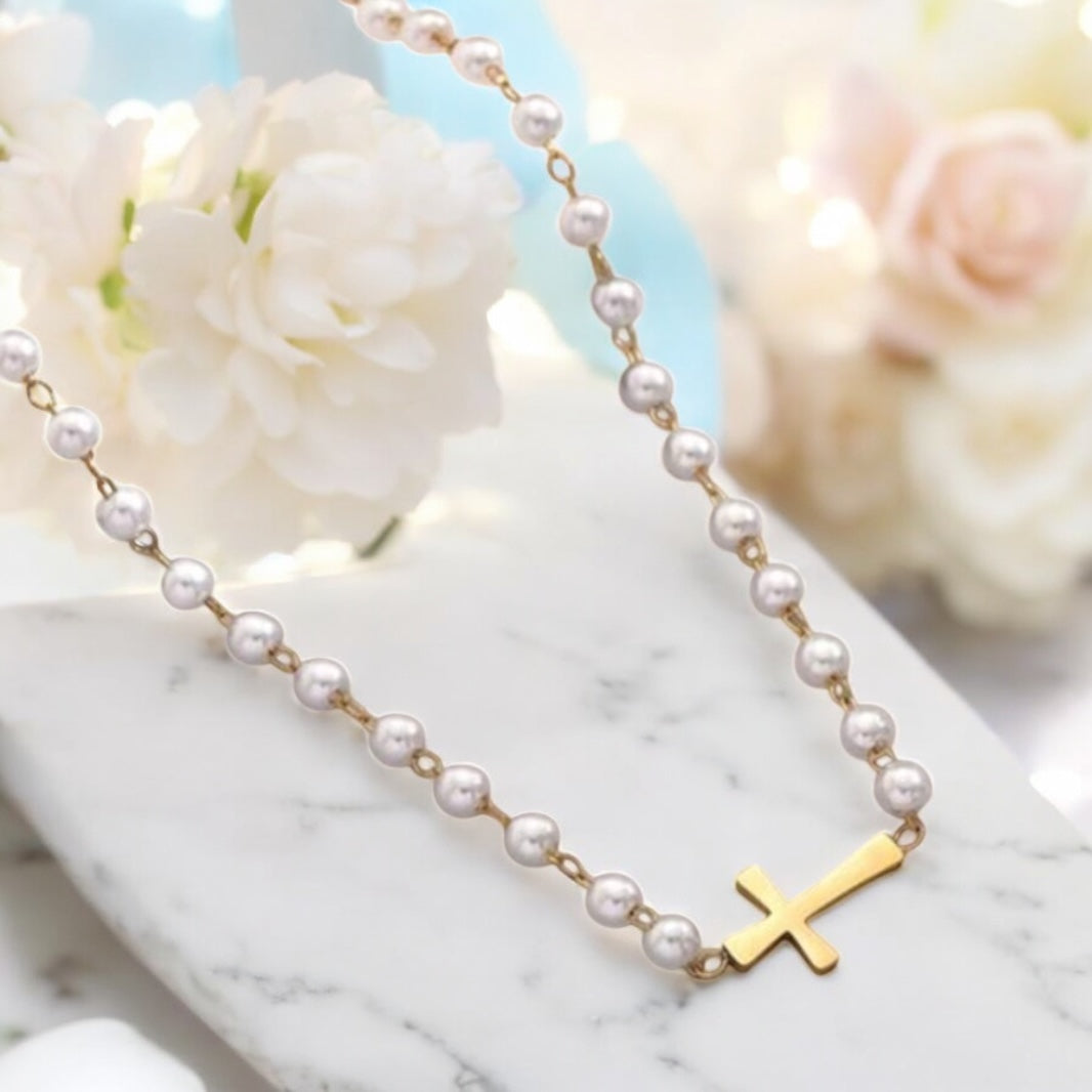 Pearl necklace with a gold cross pendant on a marble surface with flowers in the background