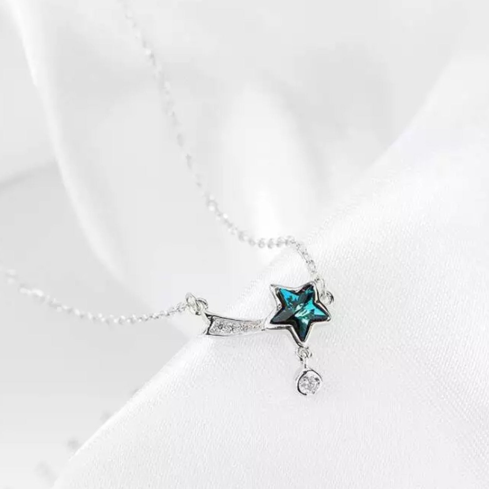 A sterling silver necklace with a blue shooting star pendant and a dangling chain, displayed on a white fabric background.