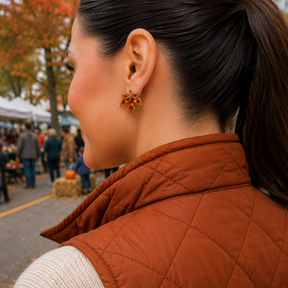 A woman in profile wearing a rust-orange quilted puffer vest and a cream knit sweater, wearing an Amber Crystal Maple Leaf Earring in an outdoor fall market setting.