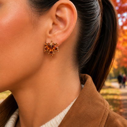 A close-up of a woman's ear wearing a Amber Crystal Maple Leaf Earrings, set against an outdoor autumn background with blurred trees.