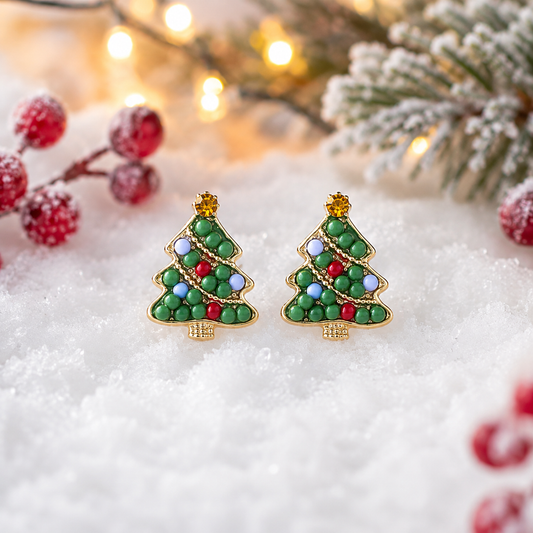 A pair of Beaded Christmas Tree Earrings decorated with green, red, blue, and white enamel and small gold beads, displayed on white faux snow with festive blurred holiday decorations in the background.