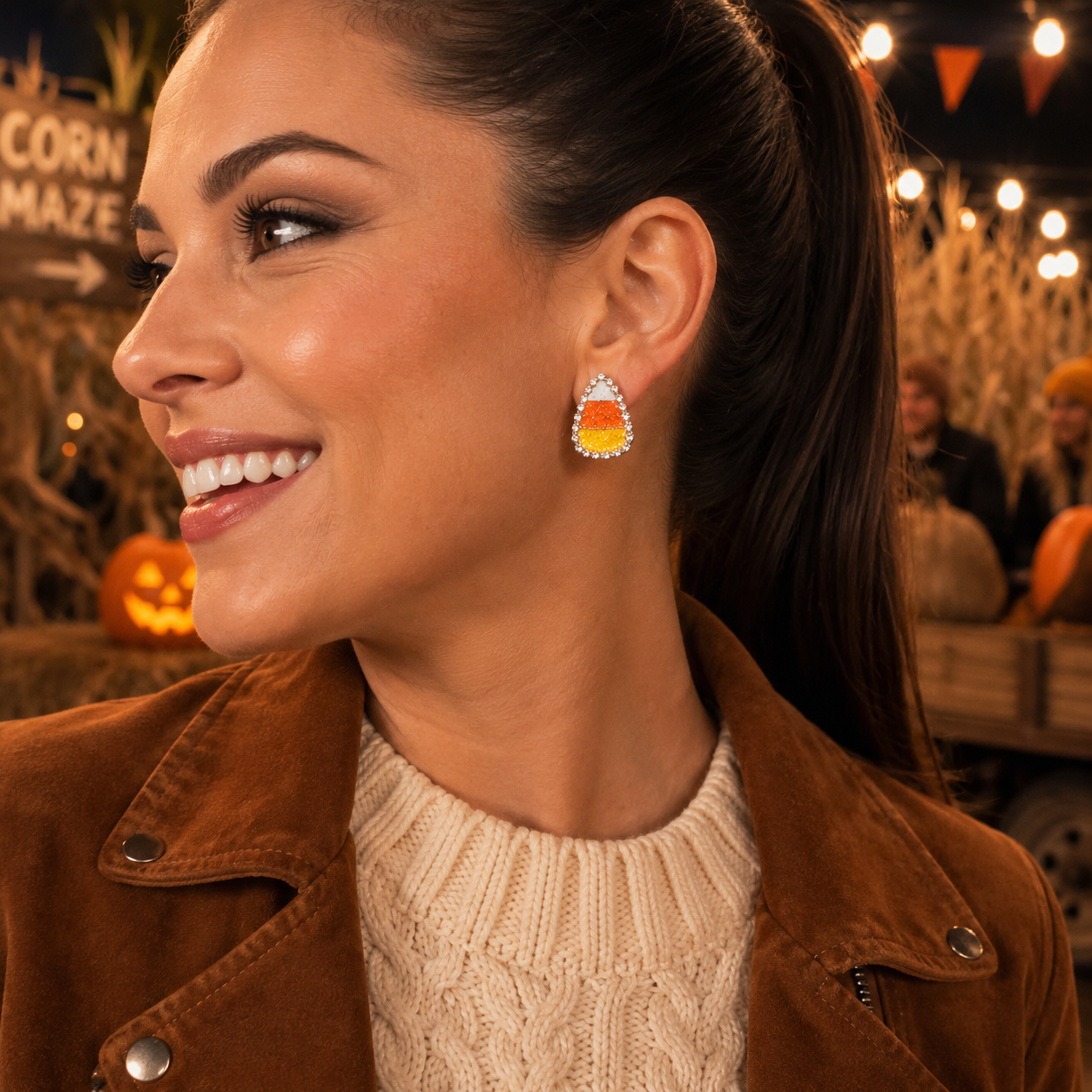 A woman in a brown suede jacket and cream cable knit sweater wears a Candy Corn Rhinestone Earrings. The background shows a warm, festive outdoor setting with pumpkins and string lights.