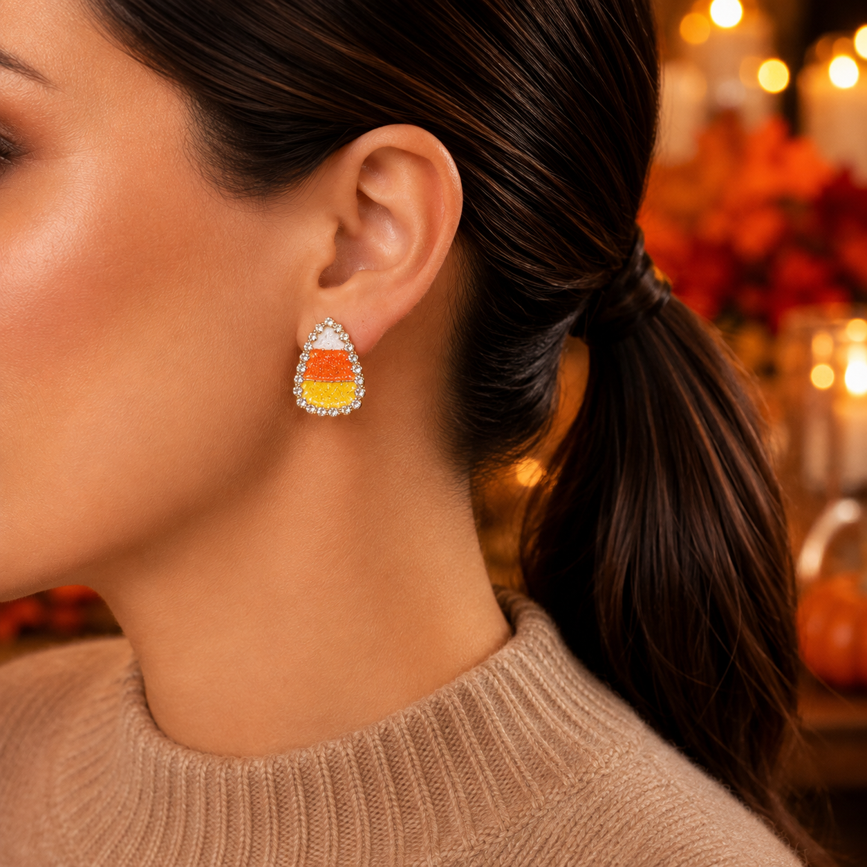 A close-up side profile of a woman wearing a Candy Corn Rhinestone Earring, set in a warm, softly blurred indoor background.