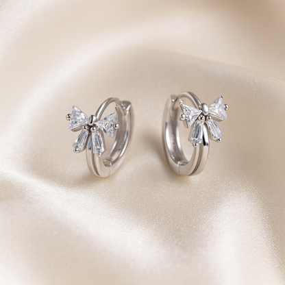 Tiny Crystal Ribbon Earrings with bow-shaped diamond accents on a beige satin background