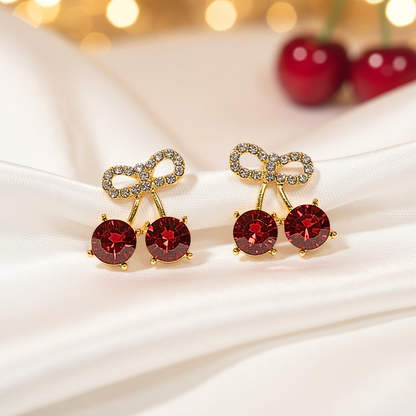 Cherry Bow Pavé Earrings with red gemstones and gold accents on a white fabric background.