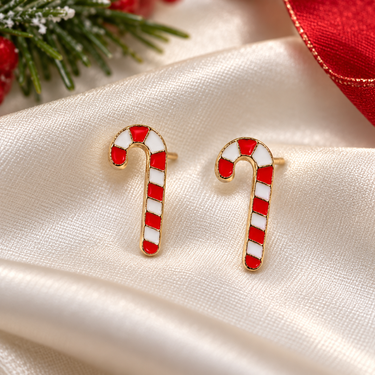 A pair of gold-tone Classic Striped Candy Cane Earrings with red and white enamel stripes, displayed on a draped cream satin fabric with festive green pine branches and a red ribbon in the background.